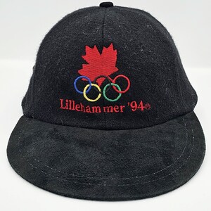 Op de afbeelding: Zwarte baseballpet met een rood esdoornblad en vijf in elkaar grijpende ringen, geborduurd met de tekst "Lillehammer '94".