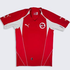 Könnte beinhalten: Ein rot-weißes Kurzarmtrikot mit V-Ausschnitt. Auf der Vorderseite befindet sich ein weißes Puma-Logo auf der rechten Brust und ein kreisförmiges Emblem des Schweizerischen Fussballverbands auf der linken Seite. Kleine Schweizer Flaggen sind an den Ärmeln.