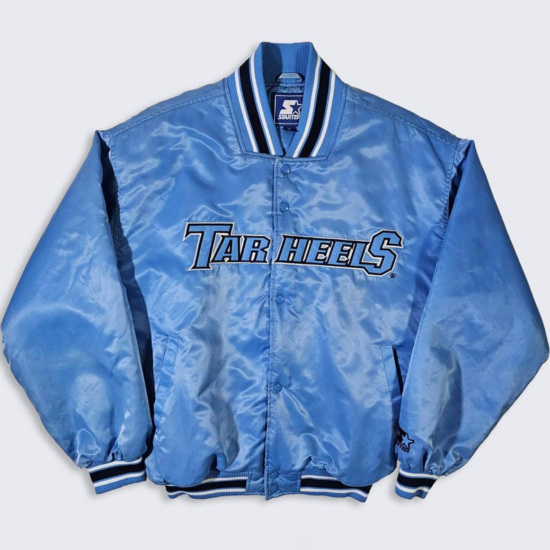 May include: Light blue satin jacket with the text "Tar Heels" across the chest. The jacket has a button-up front, a ribbed collar, cuffs, and hem with black and white stripes. The Starter logo is visible on the collar.