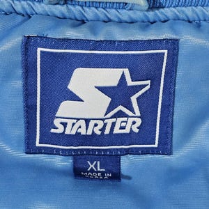 May include: Close-up of a blue Starter jacket label. The label is a dark blue rectangle with a white border. The Starter logo is in white, with a large 'S' and a star. The label also has an XL size tag and says 'Made in Korea'.