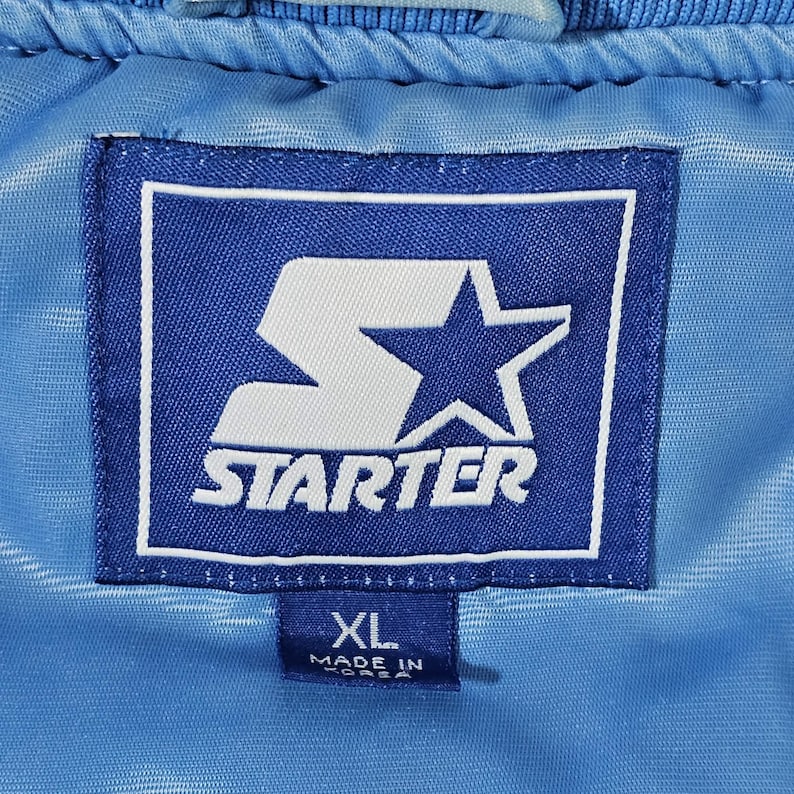 May include: Close-up of a blue Starter jacket label. The label is a dark blue rectangle with a white border. The Starter logo is in white, with a large 'S' and a star. The label also has an XL size tag and says 'Made in Korea'.