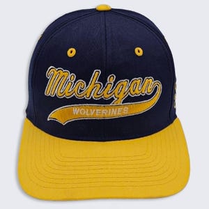 May include: A navy blue baseball cap with a yellow brim and a yellow button on top. The front features embroidered yellow text that reads Michigan with the word Wolverines in white inside a yellow swoosh graphic across the center of the cap.