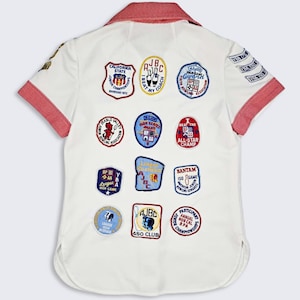 May include: A white short-sleeved shirt with a red collar and sleeve trim, featuring numerous embroidered bowling patches on the back. The patches include text such as California State, AJBC, I Beat My Coach, All-Star Champ, and Bantam 150 Game.