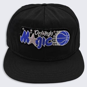 May include: A black baseball cap featuring an embroidered patch on the front. The patch displays the text Orlando Magic in blue and silver, accented with small silver stars and a blue basketball graphic on the right side of the lettering.