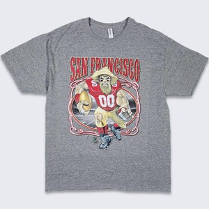May include: Gray t-shirt with a cartoon illustration of a football player wearing a red and gold uniform. The text "SAN FRANCISCO" is printed above the illustration.