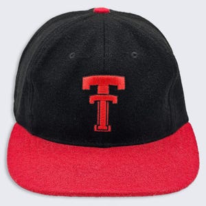 May include: A black baseball cap with a red flat brim and a red embroidered letter T logo on the front. The cap features a structured crown with a small red button at the top center and matching red stitching on the brim and the embroidered letter.