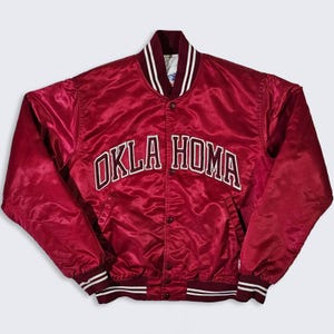May include: A maroon satin varsity jacket featuring the text OKLA HOMA arched across the chest in white and maroon lettering. The jacket includes a snap-button front closure, a ribbed collar, and ribbed cuffs with white stripes on a maroon base.