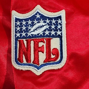 May include: A white and blue embroidered patch with the word "NFL" in red lettering. The patch has a shield shape with stars around the border.