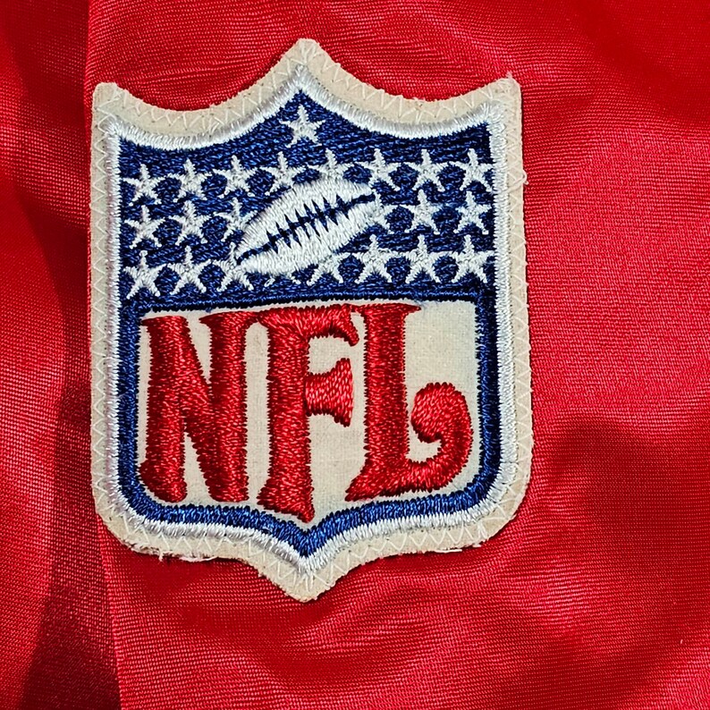 May include: A white and blue embroidered patch with the word "NFL" in red lettering. The patch has a shield shape with stars around the border.