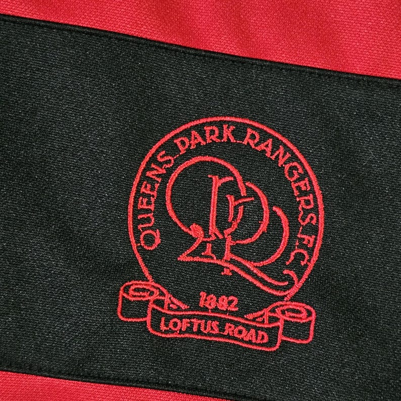 K&ouml;nnte beinhalten: Nahaufnahme eines schwarz-roten Stoffes mit einem rot gestickten Queens Park Rangers FC-Logo. Das Logo enth&auml;lt den Text "Queens Park Rangers FC", "1882" und "Loftus Road".