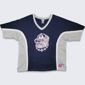 Puede incluir: Una camiseta Champion azul marino y gris con un bulldog blanco mascota que lleva una gorra de béisbol con una "G" en ella. La camiseta tiene cuello en V y mangas cortas.