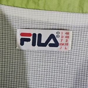 May include: Close-up of a Fila clothing label. The label is rectangular with the brand name in navy blue and red. The background is a white and gray checkered pattern, with a green border at the top.
