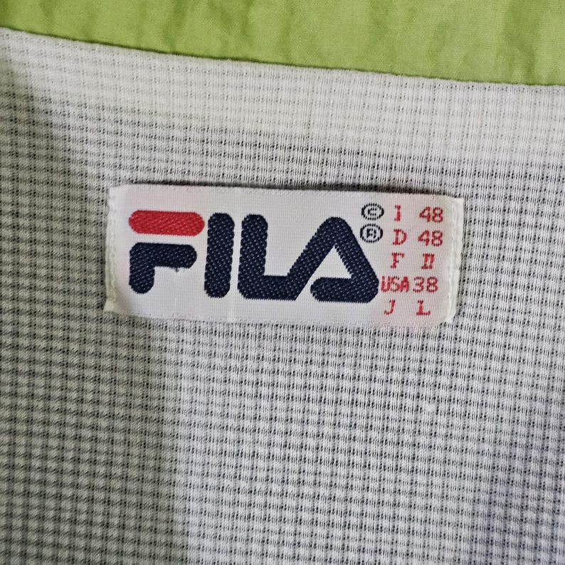 May include: Close-up of a Fila clothing label. The label is rectangular with the brand name in navy blue and red. The background is a white and gray checkered pattern, with a green border at the top.