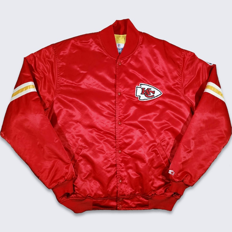 May include: A red satin bomber jacket with a white and gold striped sleeve. The jacket has a snap closure and a large embroidered patch on the left chest with the letters "KC" in a red arrowhead.
