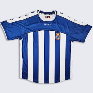 Puede incluir: Una camiseta de fútbol azul y blanca a rayas con el logotipo de Kelme y un escudo de equipo. La camiseta tiene mangas cortas y cuello redondo.