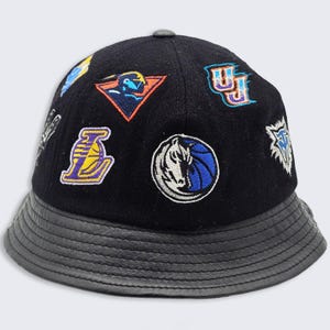 May include: A black bucket hat featuring a textured brim and multiple embroidered sports team patches across the crown. The patches include a purple and yellow basketball logo, a blue and white horse head, and various other colorful graphic emblems.