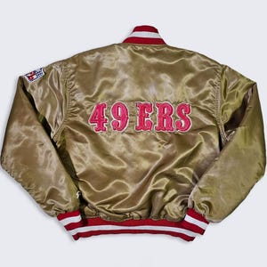 May include: A gold satin jacket with red and white accents. The back of the jacket features the embroidered words "49 ERS" in red. The jacket has a red and white striped collar, cuffs, and waistband. A patch is visible on the left sleeve.