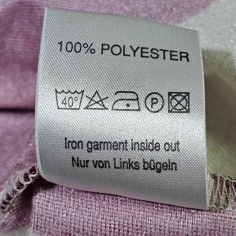 Puede incluir: Una etiqueta de cuidado blanca con texto negro que dice "100% POLYESTER". La etiqueta tambi&eacute;n incluye un conjunto de instrucciones de cuidado: lavar a m&aacute;quina a 40 grados Celsius, no blanquear, planchar a baja temperatura, no limpiar en seco y planchar del rev&eacute;s. El texto "Iron garment inside out Nur von Links b&uuml;geln" tambi&eacute;n est&aacute; incluido.