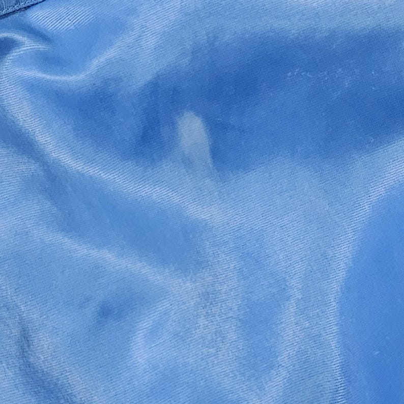 May include: Close-up of a light blue satin fabric with a white stain. The fabric has a smooth, shiny texture and is slightly wrinkled. The lighting creates highlights and shadows, emphasizing the fabric's folds and sheen.