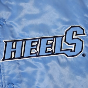 May include: Light blue satin jacket with the word "HEELS" in a bold, two-tone design. The lettering is in a darker shade of blue with a white outline. The jacket has a snap closure and a shiny finish.