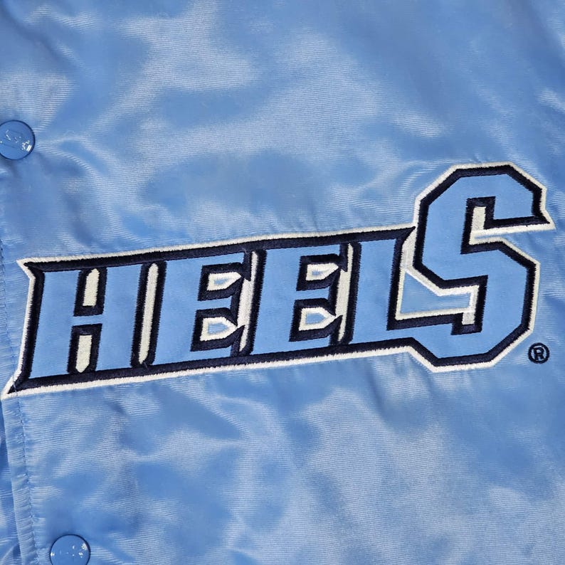 May include: Light blue satin jacket with the word "HEELS" in a bold, two-tone design. The lettering is in a darker shade of blue with a white outline. The jacket has a snap closure and a shiny finish.