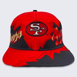 May include: Red and black baseball cap with a San Francisco 49ers logo and a graphic design of jagged lines and the word "49ers" in yellow.