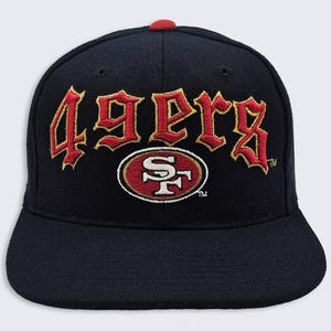 May include: A navy blue baseball cap with a red top button. The front features the word "49ers" in red and gold embroidery above a red and gold oval logo with a white "SF" design.