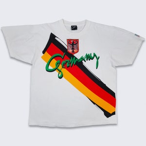 May include: White t-shirt with a diagonal graphic of the German flag and the word "Germany" in green lettering. The graphic also includes the German coat of arms.