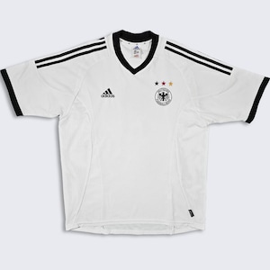May include: White soccer jersey with black Adidas logo and three stars above the German national team crest. The jersey has black stripes on the shoulders and black trim around the collar.