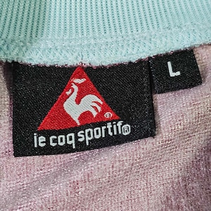 Puede incluir: Una etiqueta negra y roja con un logotipo de gallo y el texto "le coq sportif" en blanco. La etiqueta est&aacute; unida a una camisa azul claro y rosa con una etiqueta de talla "L".
