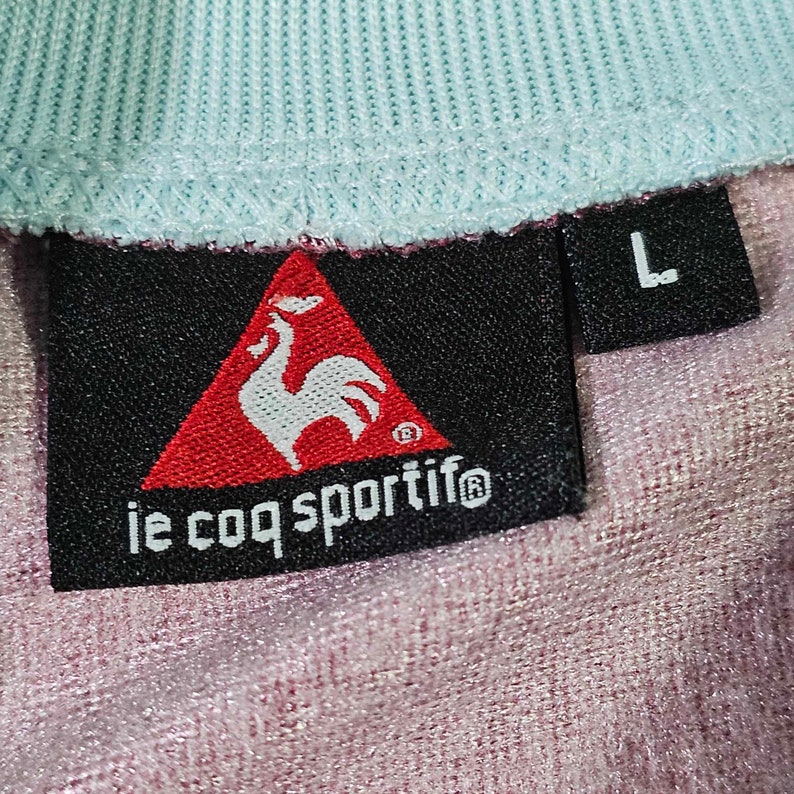 Puede incluir: Una etiqueta negra y roja con un logotipo de gallo y el texto "le coq sportif" en blanco. La etiqueta est&aacute; unida a una camisa azul claro y rosa con una etiqueta de talla "L".