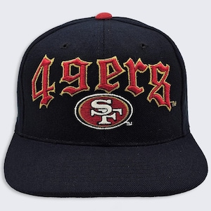 Op de afbeelding: Een marineblauwe honkbalpet met de tekst 49ERS geborduurd in rood met gouden omlijningen aan de voorzijde. Onder de tekst staat een ovaalvormig logo van het San Francisco 49ers-team in rood, wit en goud. De pet heeft een rode knoop bovenop.