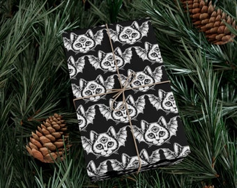May include: Black wrapping paper featuring a repeating pattern of a cat with bat wings and a third eye. The paper is tied with twine and set against a backdrop of pine needles and pine cones.
