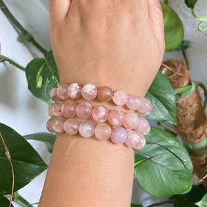 May include: A set of three pink agate beaded bracelets with pink and white patterns. The bracelets are stacked on a person's wrist.