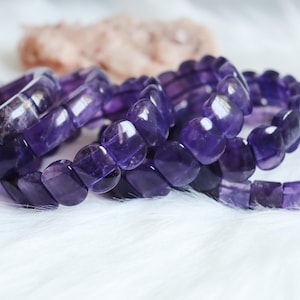 Puede incluir: Una colección de pulseras de amatista de color púrpura intenso con cuentas rectangulares pulidas. Las piedras translúcidas y facetadas muestran diversos tonos de violeta y morado oscuro, creando una superficie lisa y reflectante en las piezas de joyería.