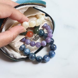 May include: A teal zippered pouch with several beaded bracelets. The bracelets feature various colors, including cream, purple, blue, and orange. The beads are round and appear to be made of stone or glass.