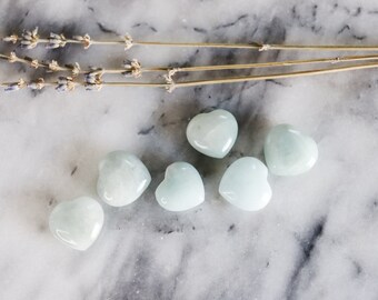 May include: Six heart-shaped, pale green stones on a white marble surface. The stones are smooth and polished.