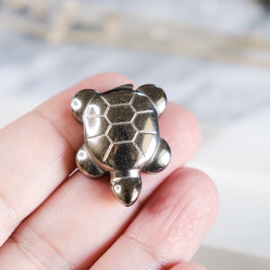 May include: A small, silver-coloured turtle figurine made of polished metal. The turtle has a detailed shell with a geometric pattern.