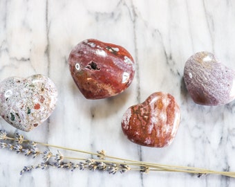 May include: Four heart-shaped stones in red, white, and purple hues are displayed on a marble surface, accompanied by lavender. The stones have varied patterns and colours, with speckled and marbled textures.