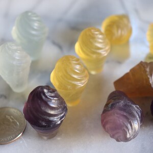 May include: A collection of small, colorful, frosted glass cupcake-shaped ornaments. The ornaments are in shades of purple, yellow, and white.