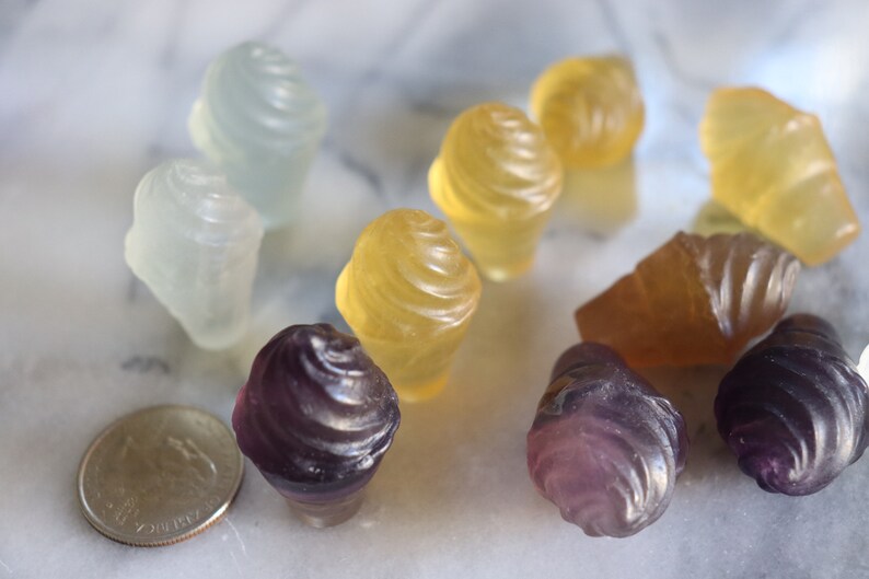 May include: A collection of small, colorful, frosted glass cupcake-shaped ornaments. The ornaments are in shades of purple, yellow, and white.
