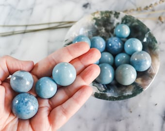 May include: A collection of smooth, light blue gemstone spheres in a hand and a bowl. The spheres are polished and have a subtle shimmer.
