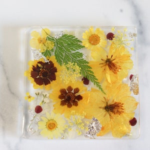 Puede incluir: Un posavasos cuadrado de resina con flores y hojas prensadas de color amarillo, marrón y verde. El posavasos tiene un acabado de resina transparente.