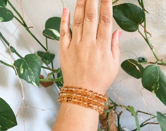 May include: A close-up of a person's wrist wearing four stacked bracelets made of small, round, amber-colored beads.