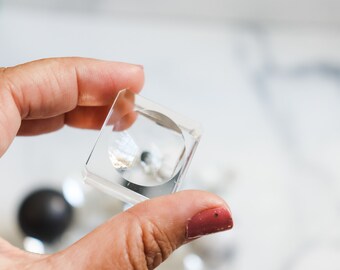 May include: A clear, square-shaped object with a circular cutout, held in a hand. The object appears to be made of transparent acrylic or glass. The background is blurred, with other objects visible.