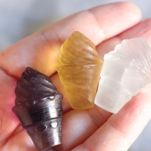 May include: Three small ice cream cone shaped candies in a hand. The candies are brown, yellow, and white.