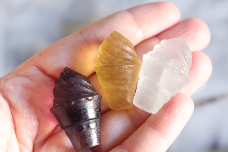 May include: Three small ice cream cone shaped candies in a hand. The candies are brown, yellow, and white.