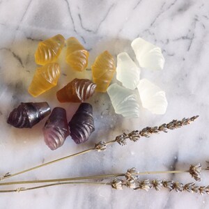 May include: Assortment of handmade, colorful, and translucent ice cream cone shaped bath bombs. The bath bombs are arranged on a white marble surface with dried lavender sprigs.