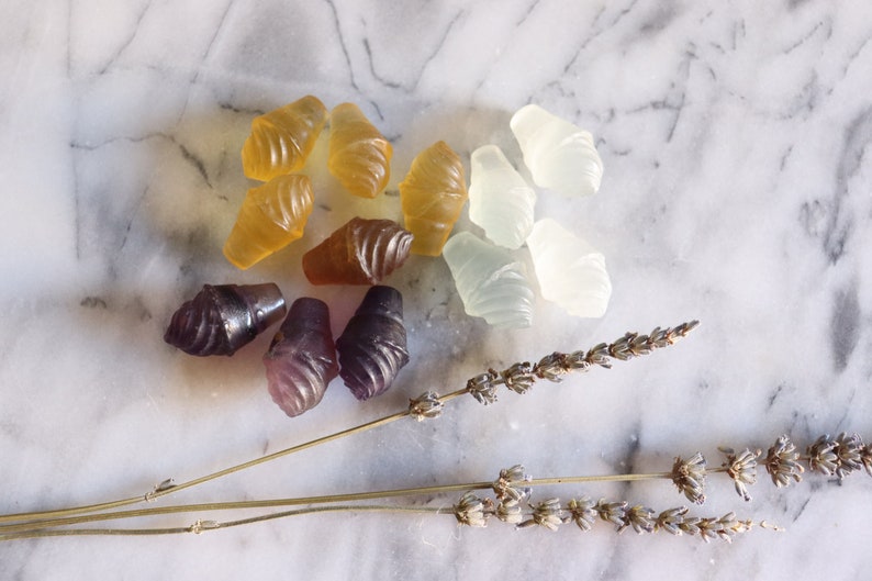 May include: Assortment of handmade, colorful, and translucent ice cream cone shaped bath bombs. The bath bombs are arranged on a white marble surface with dried lavender sprigs.
