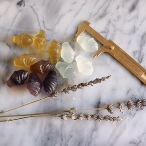 May include: A collection of colorful, translucent, shell-shaped candies on a marble surface. The candies are arranged in a cluster, with some overlapping. A gold-colored caliper is placed to the right of the candies, and a dried lavender sprig is placed below the candies.